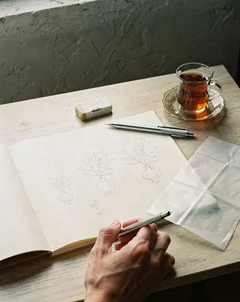 A sketchbook with one design drawn in three sizes, next to a glass of tea